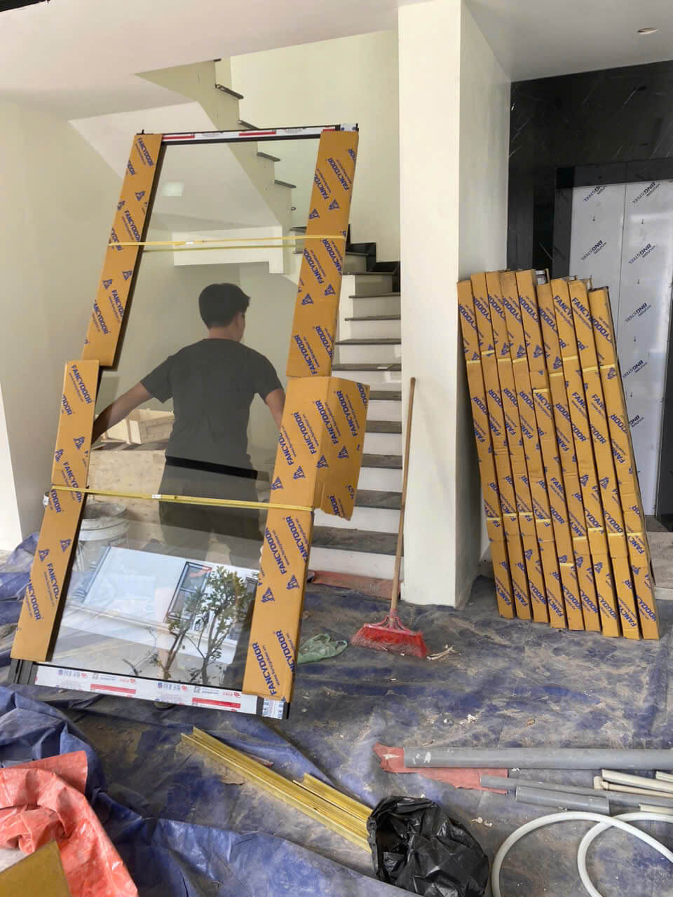 Cửa kính cường lực Newdoor đang lắp đặt trong nhà, kèm nhiều khung kính Newdoor xếp cạnh cầu thang